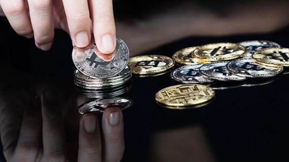 A person picking up a Bitcoin coin from a pile of other coins on a dark surface.